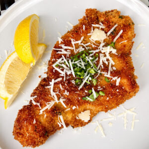 A breaded and fried cutlet topped with grated cheese and chopped herbs, served on a white plate with two lemon wedges on the side.