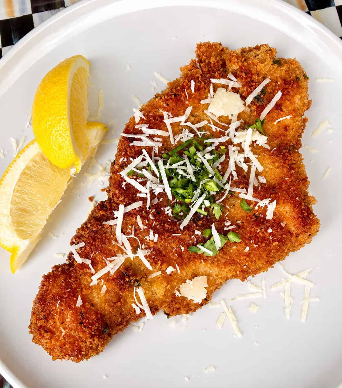 A breaded and fried cutlet topped with grated cheese and chopped herbs, served on a white plate with two lemon wedges on the side.