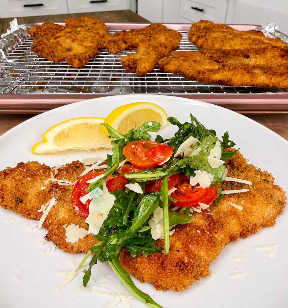 A plate of breaded and fried chicken cutlet topped with arugula, cherry tomatoes, shaved parmesan, and a lemon wedge, with more cutlets cooling on a rack in the background.