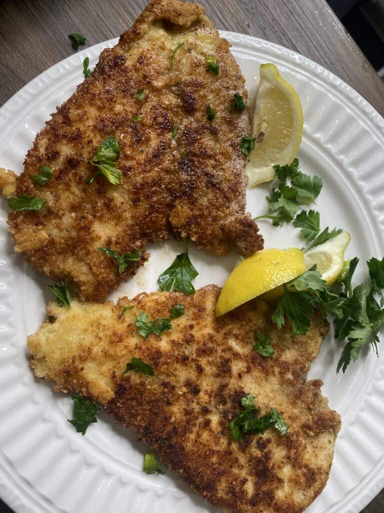 Two pieces of breaded and pan-fried schnitzel on a white plate, garnished with fresh parsley and served with two lemon wedges.