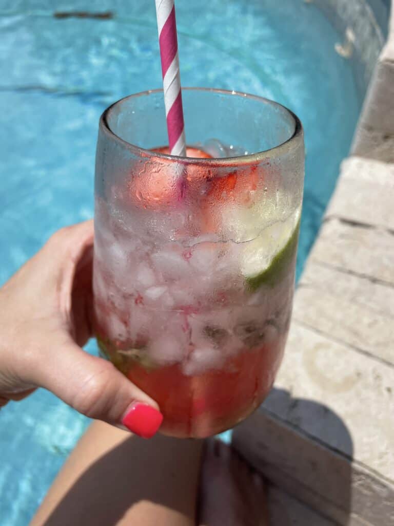 A hand with bright pink nail polish holds a glass of iced fruit drink with lime and strawberry slices, a pink and white striped straw, next to the edge of a swimming pool.