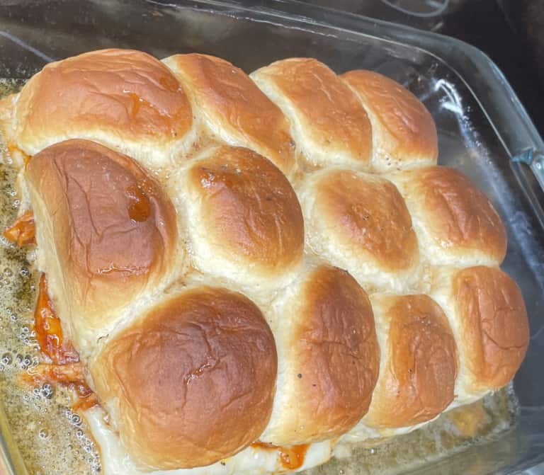 A glass baking dish filled with golden-brown baked slider sandwiches, with melted cheese and some filling visible at the edges. Butter sauce pools around the bottom of the buns.