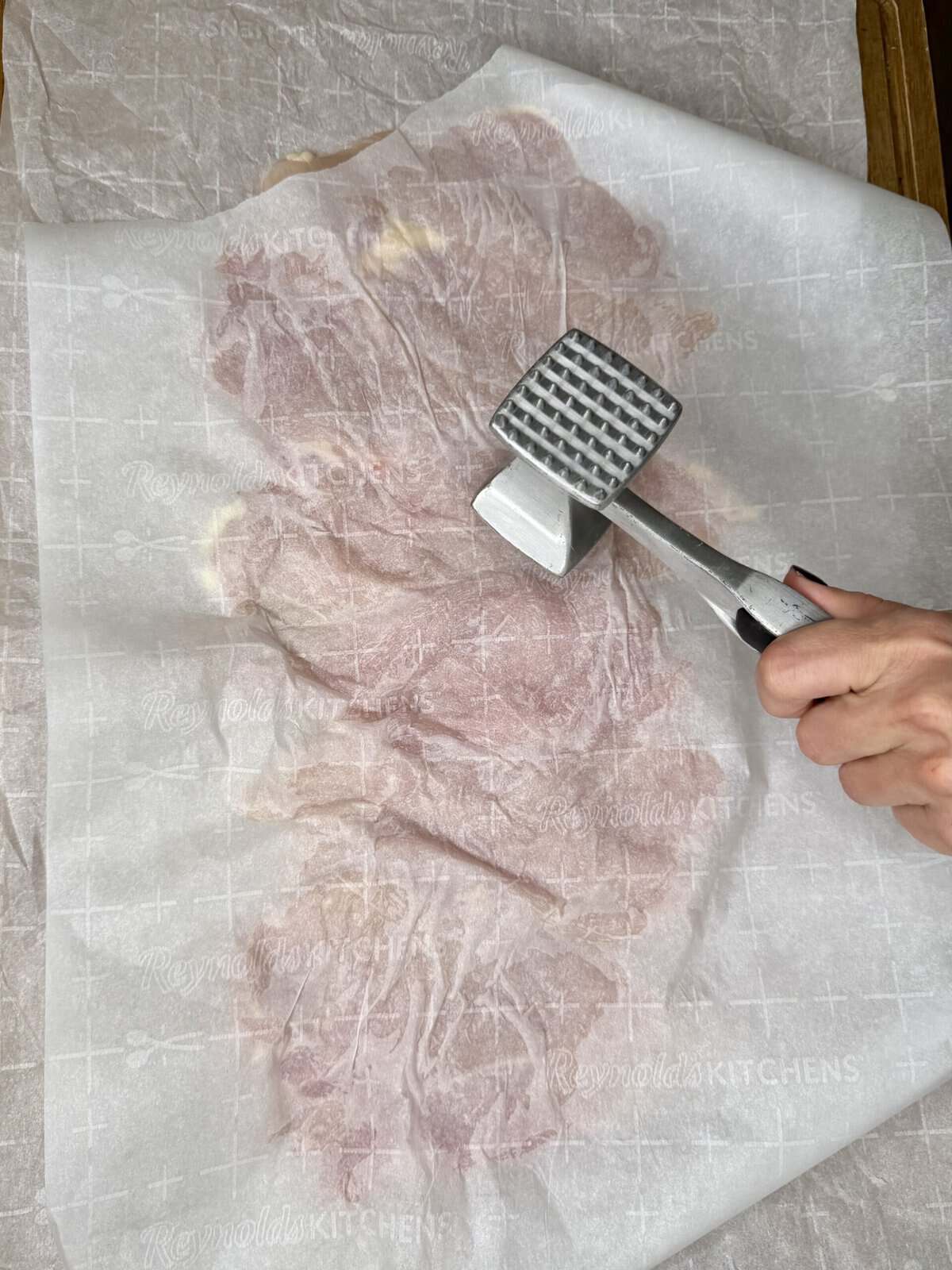 A hand uses a metal meat tenderizer to pound a piece of raw meat covered with parchment paper on a wooden surface.