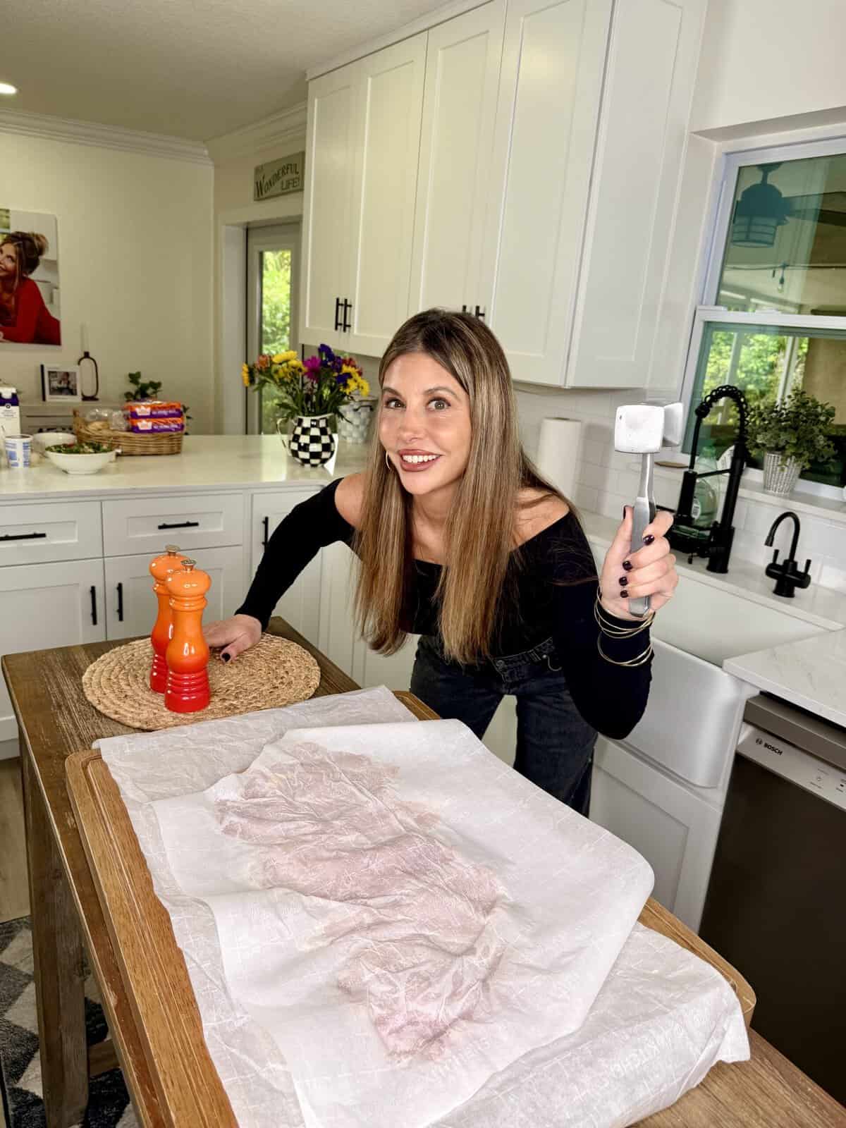 A woman in a kitchen smiles at the camera while holding a meat tenderizer over a table covered with a white cloth. A red pepper grinder and a woven placemat are also on the table. White cabinets and a farmhouse sink are in the background.