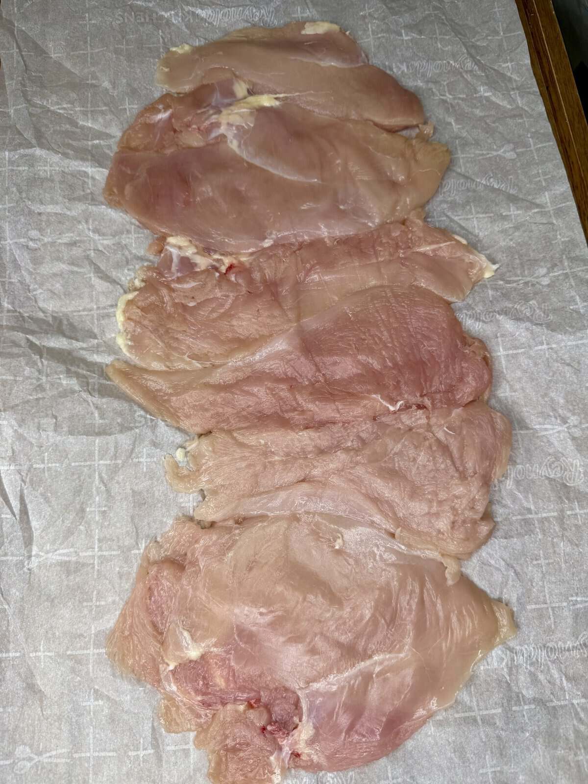 Five raw, thinly sliced pieces of chicken breast are laid out in a row on a sheet of parchment paper. The pieces vary slightly in shape and size.