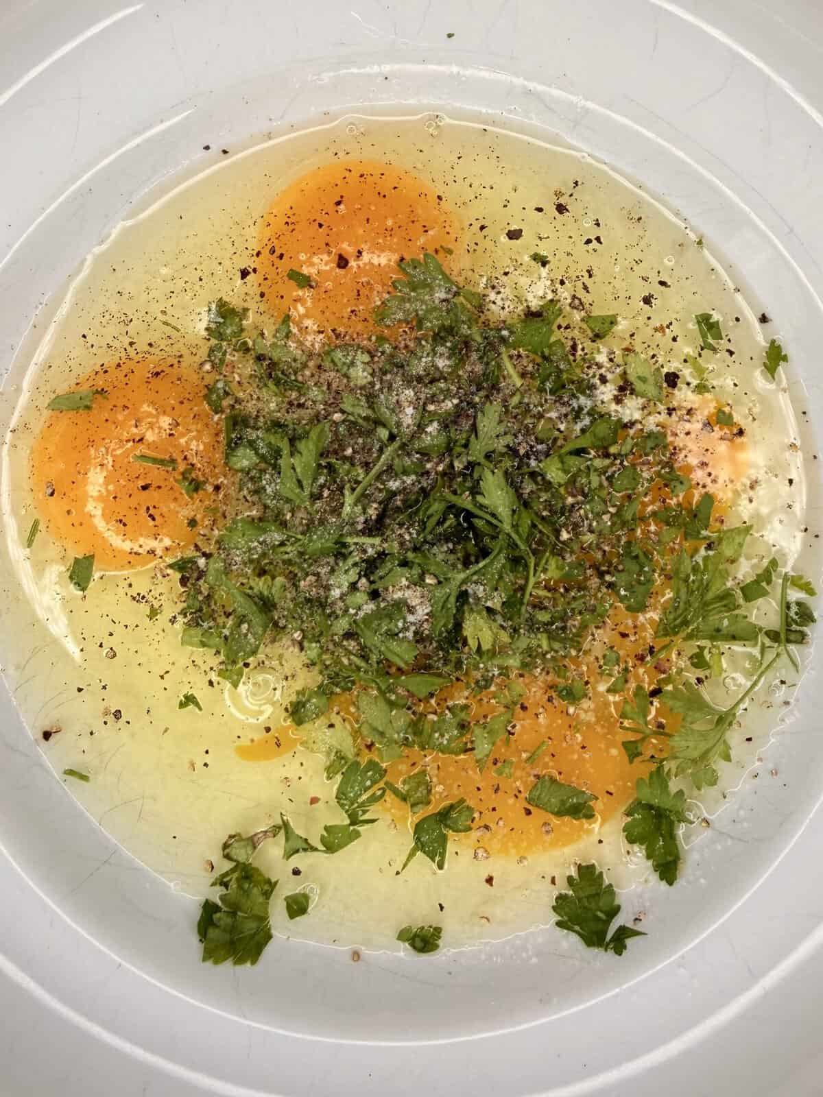 A close-up of three cracked eggs in a white bowl, topped with chopped herbs, salt, and freshly ground black pepper.