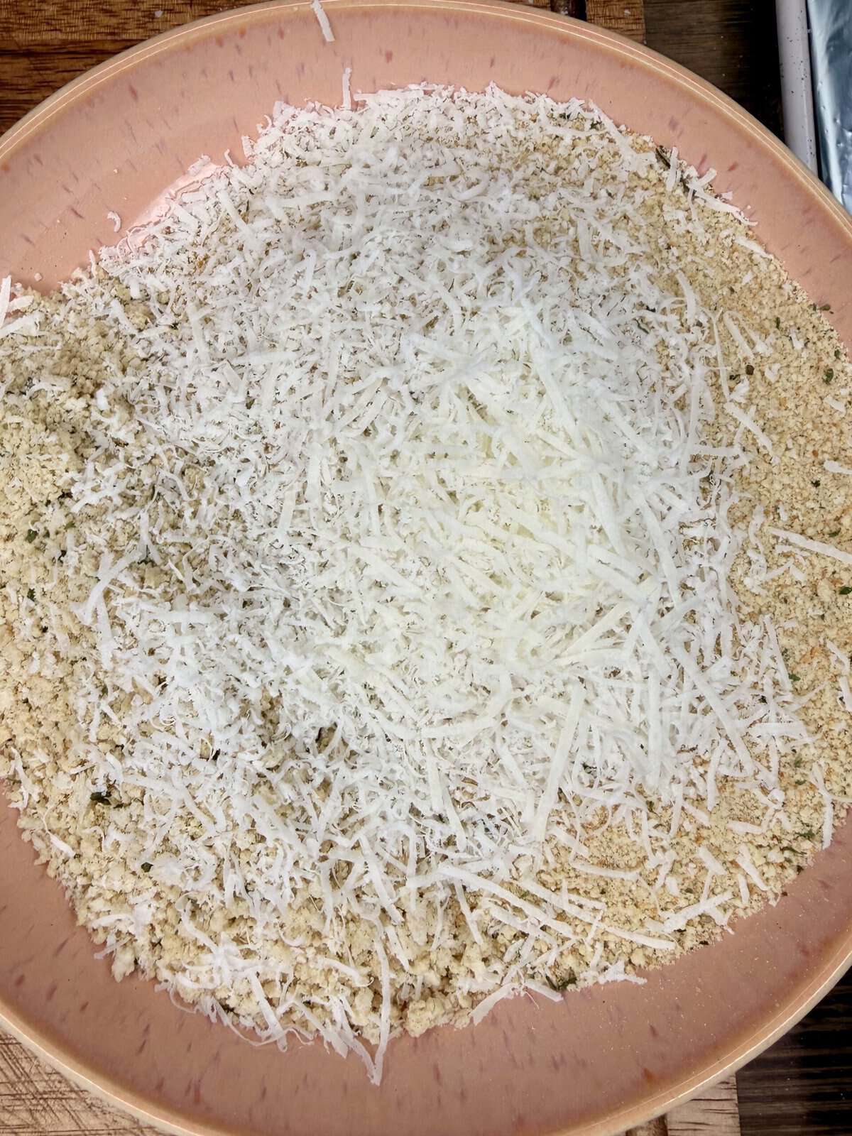 A close-up of a pink bowl filled with a mixture of breadcrumbs and finely grated white cheese, likely Parmesan, on a wooden surface.