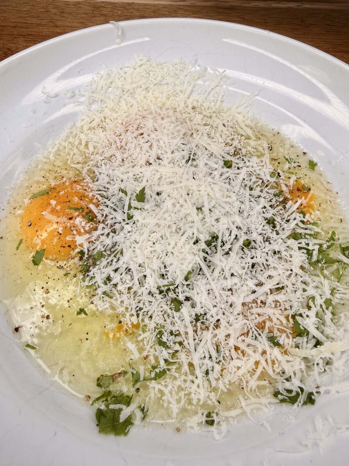 Two raw eggs in a white bowl, topped with finely grated cheese, chopped herbs, and black pepper, ready to be mixed for cooking.