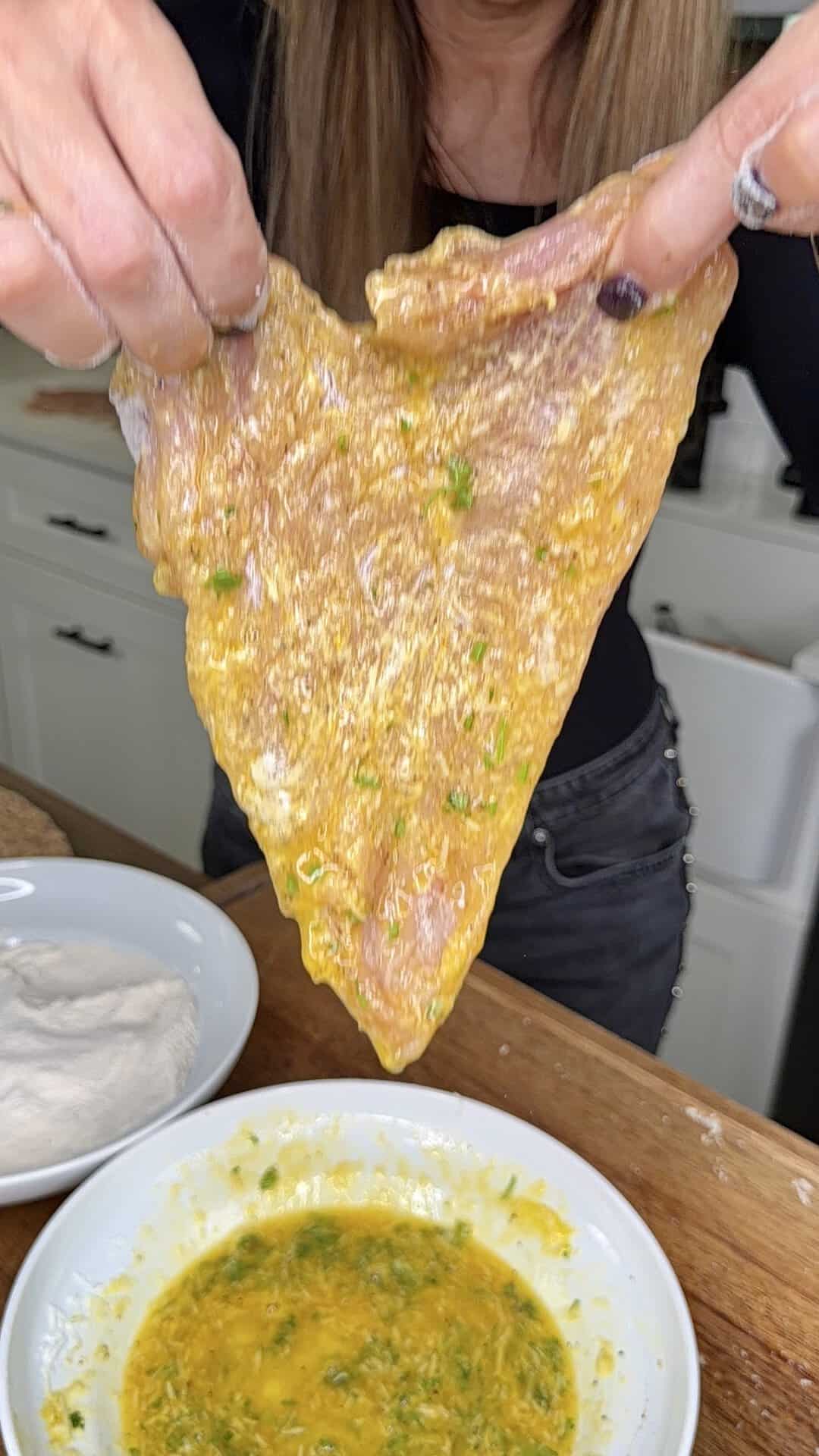 A person holds up a piece of raw, seasoned meat coated in an egg and herb mixture over a bowl, preparing to bread it in a kitchen.