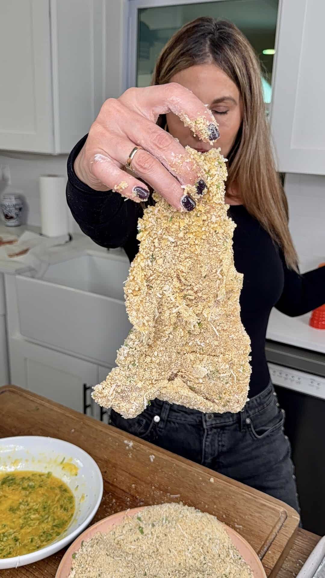 A woman in a kitchen holds up a breaded piece of meat or cutlet between her fingers, with bowls of egg wash and breadcrumbs on the counter in front of her.
