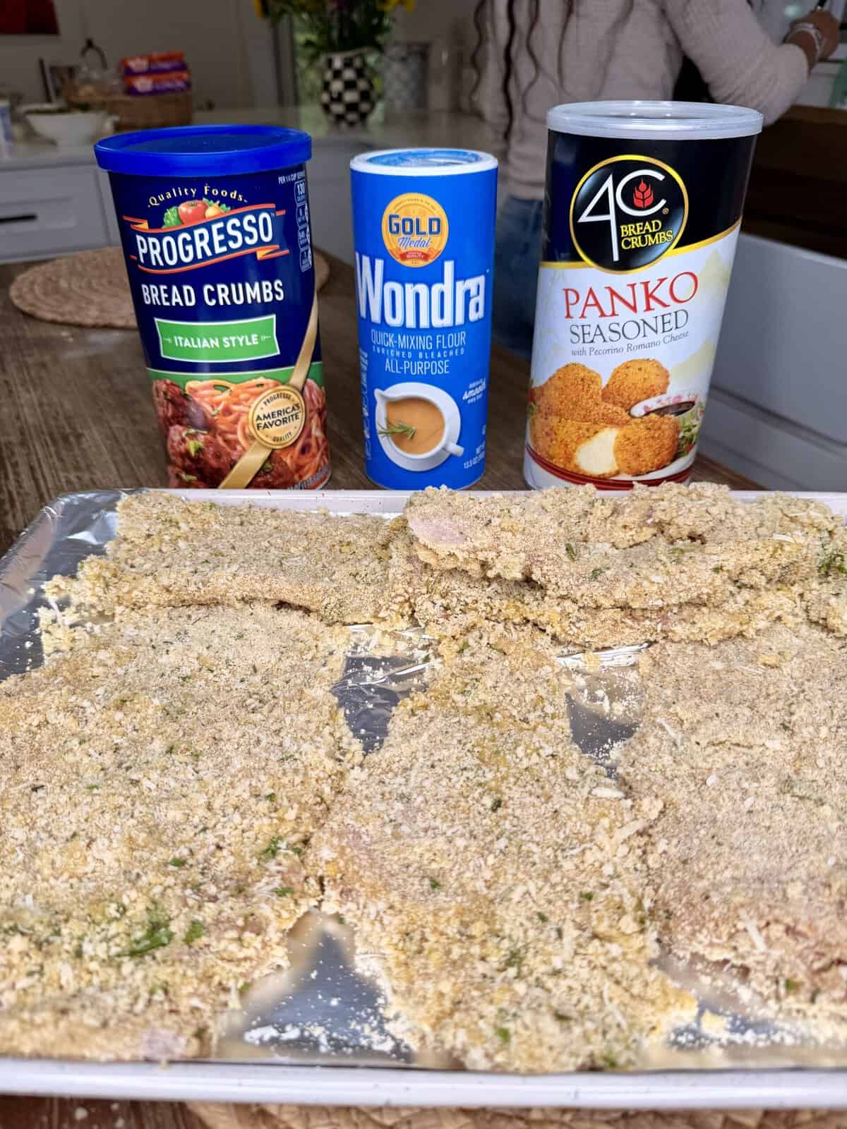 A baking tray lined with breaded cutlets sits on a counter. Behind it are cans of Progresso Italian bread crumbs, Gold Medal Wondra flour, and 4C seasoned panko bread crumbs.
