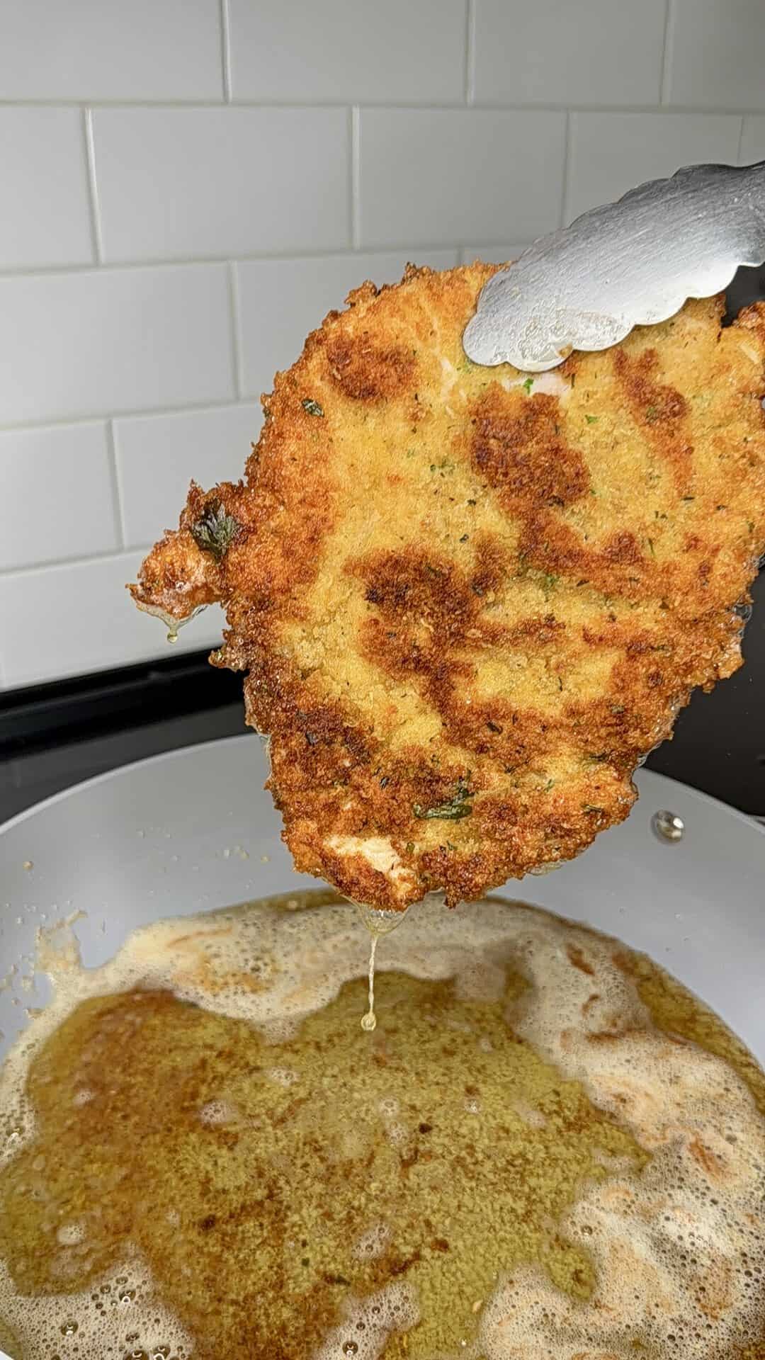 A close-up of metal tongs holding a golden-brown, breaded and fried cutlet over a frying pan filled with bubbling oil, with white subway tile backsplash in the background.