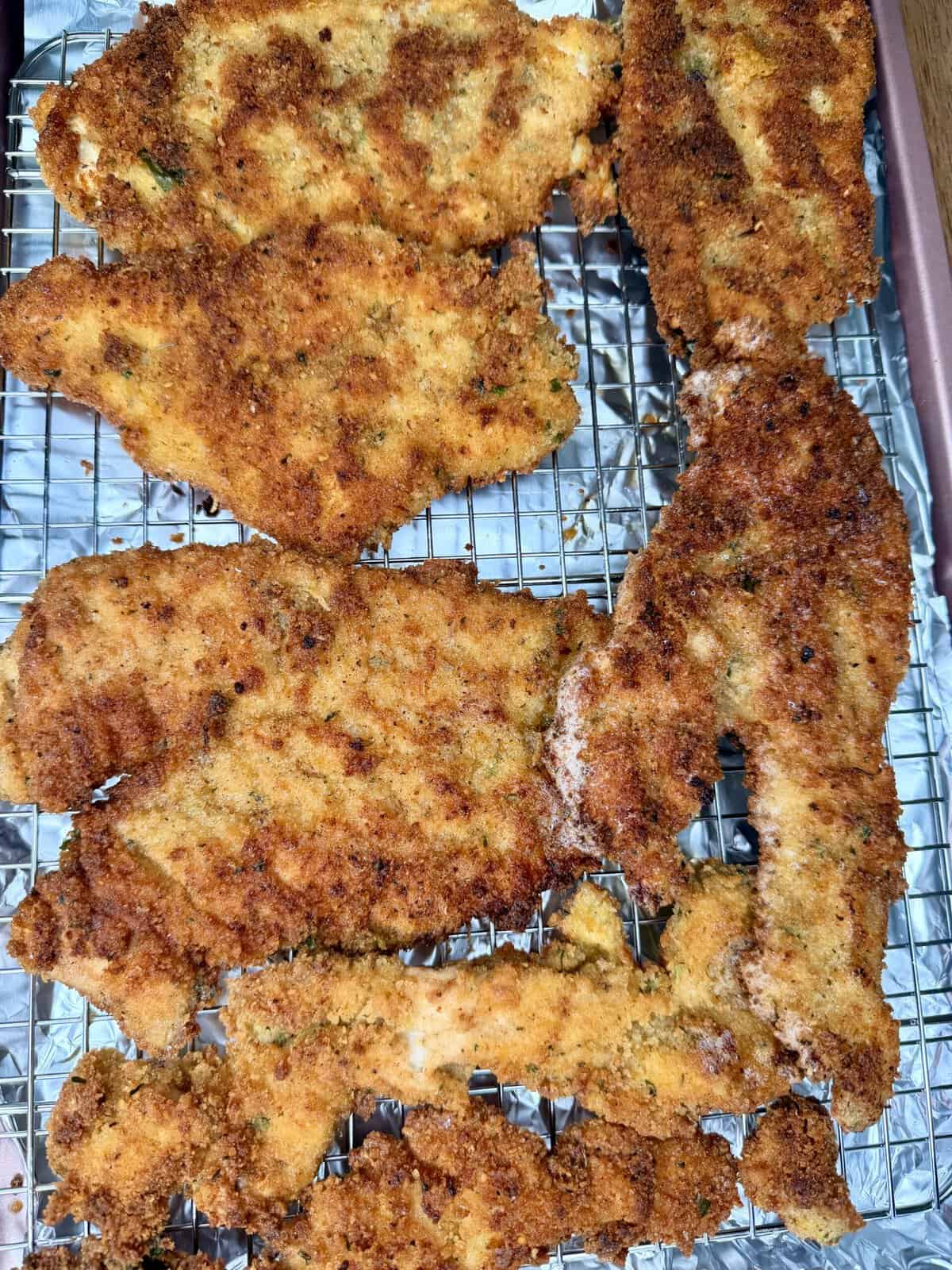 Six pieces of golden brown, crispy breaded chicken cutlets are cooling on a wire rack placed over a baking sheet. The breading appears evenly cooked and crunchy.