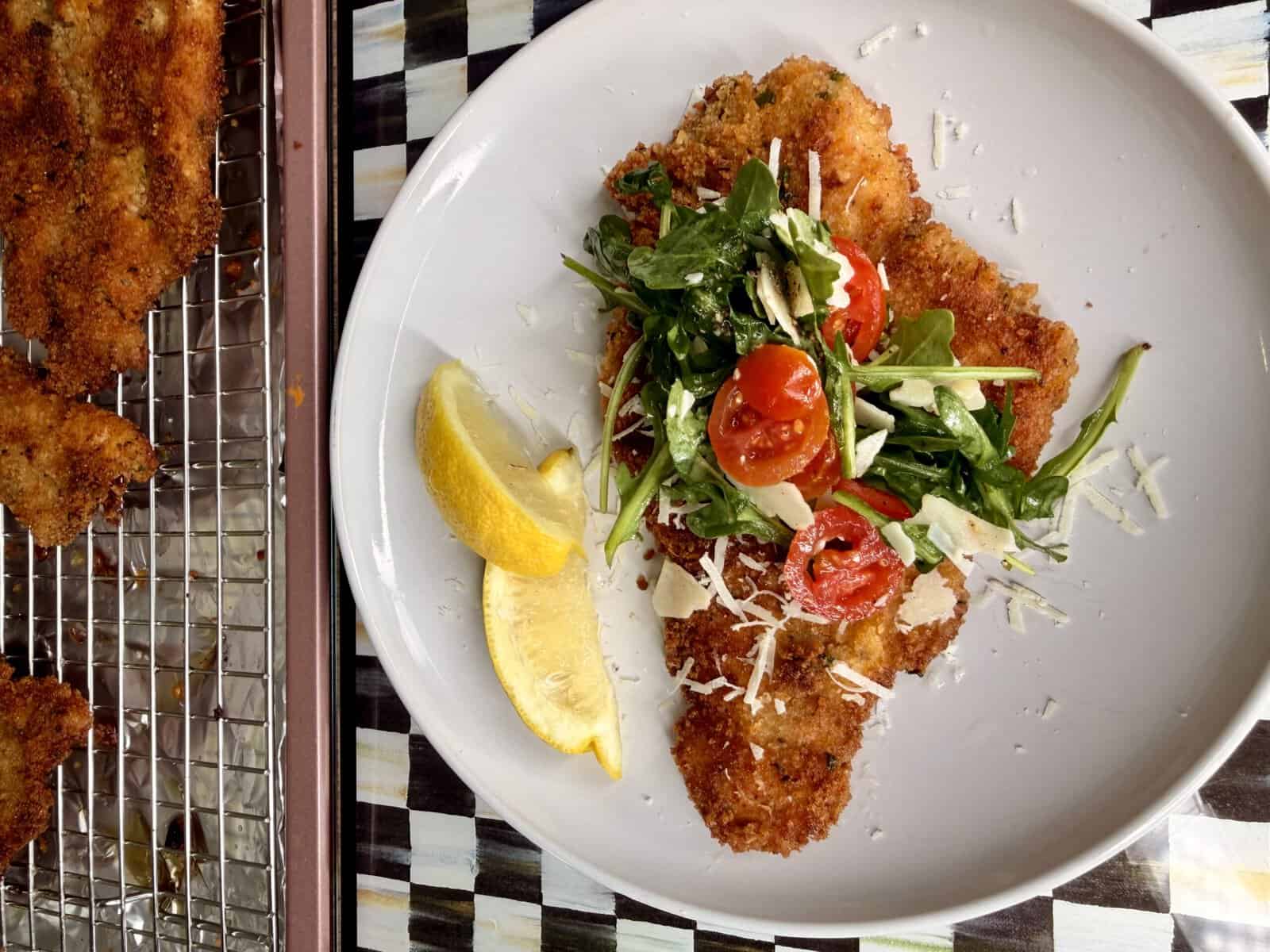 A white plate with breaded, fried cutlet topped with arugula, cherry tomatoes, and shredded cheese, garnished with lemon wedges. More cutlets are on a wire rack next to the plate.