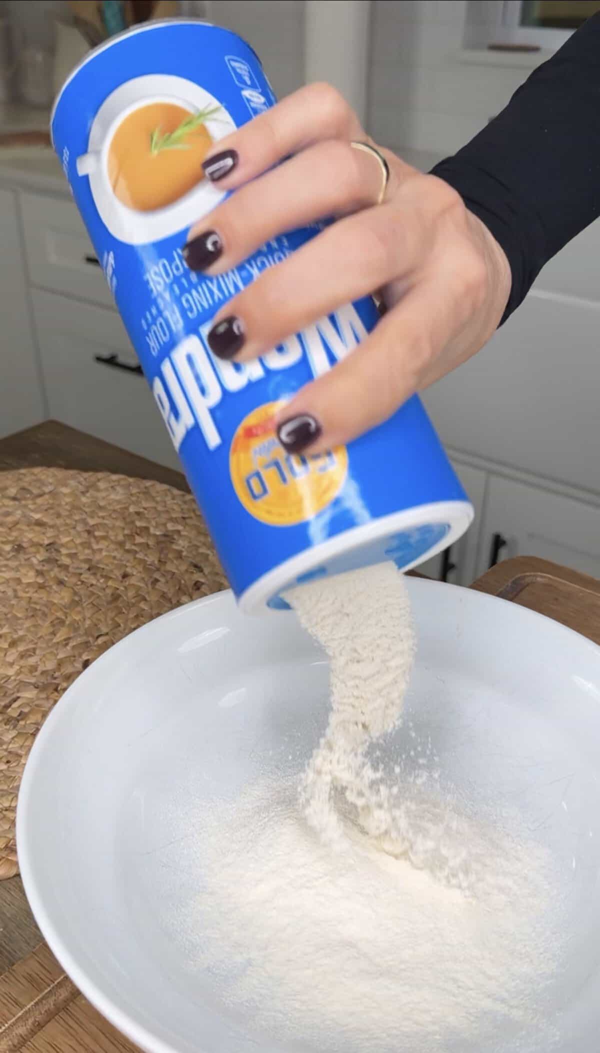 A hand pours salt from a blue Morton salt container into a white bowl on a kitchen counter.