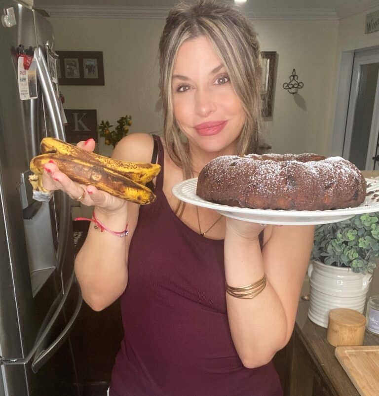 A woman in a maroon tank top stands in a kitchen, smiling and holding ripe bananas in one hand and a chocolate bundt cake dusted with powdered sugar in the other.