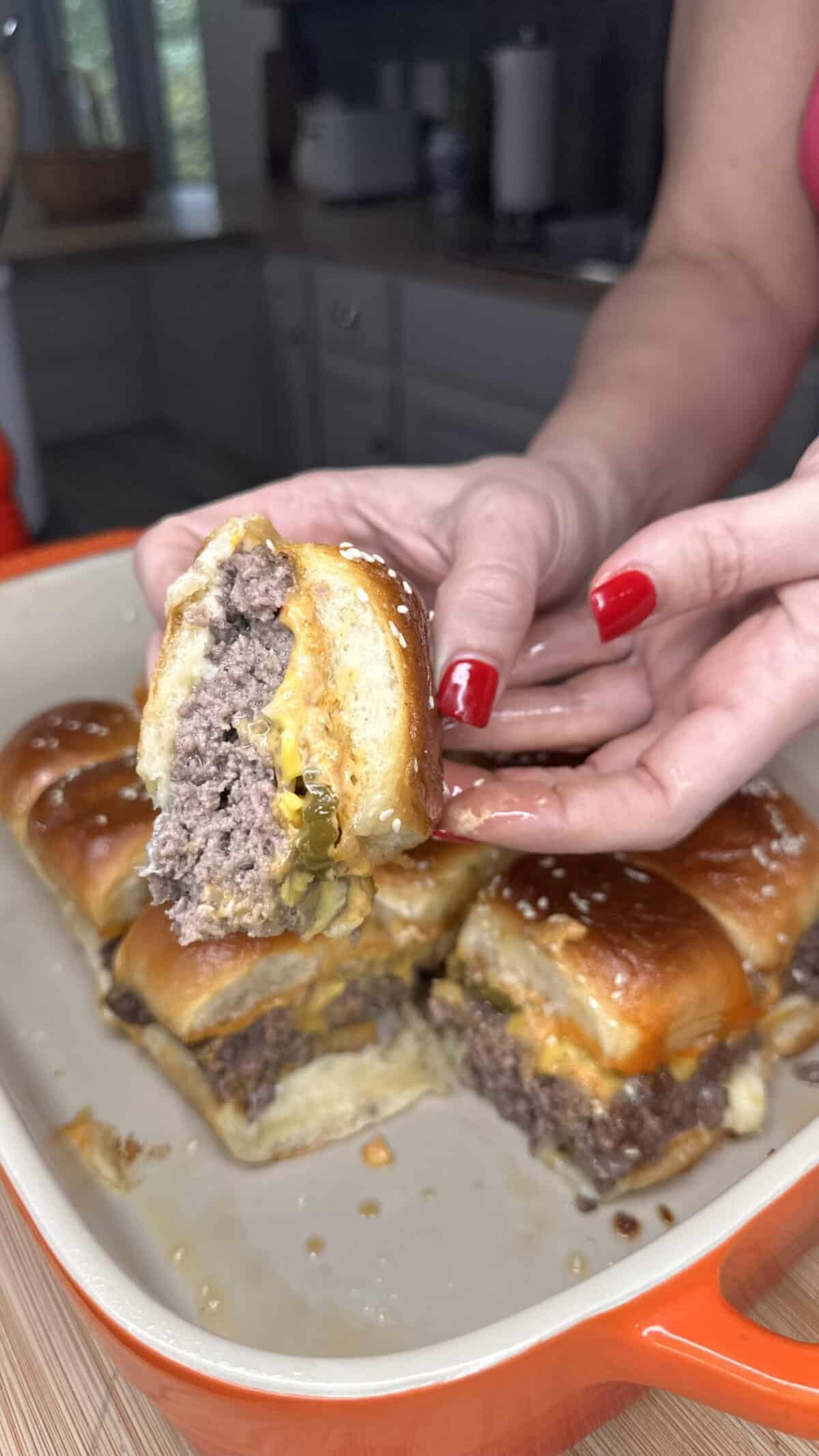 A person with red nails holds a cheeseburger slider with a beef patty and melted cheese over a baking dish filled with more sliders in a kitchen setting.