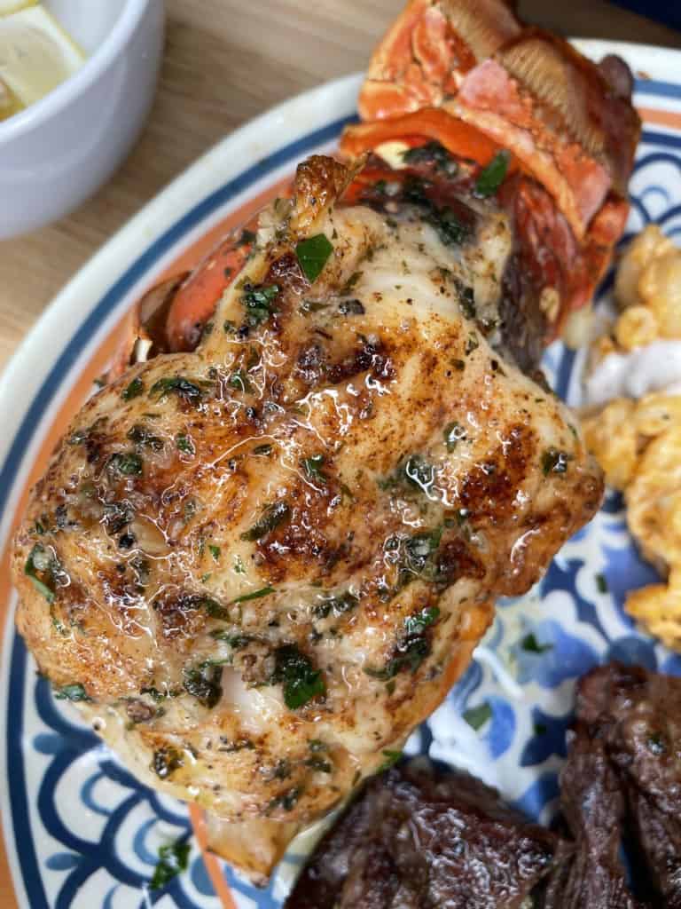 Close-up of a grilled lobster tail seasoned with herbs and spices on a decorative plate, with side dishes including scrambled eggs and pieces of grilled meat visible nearby.
