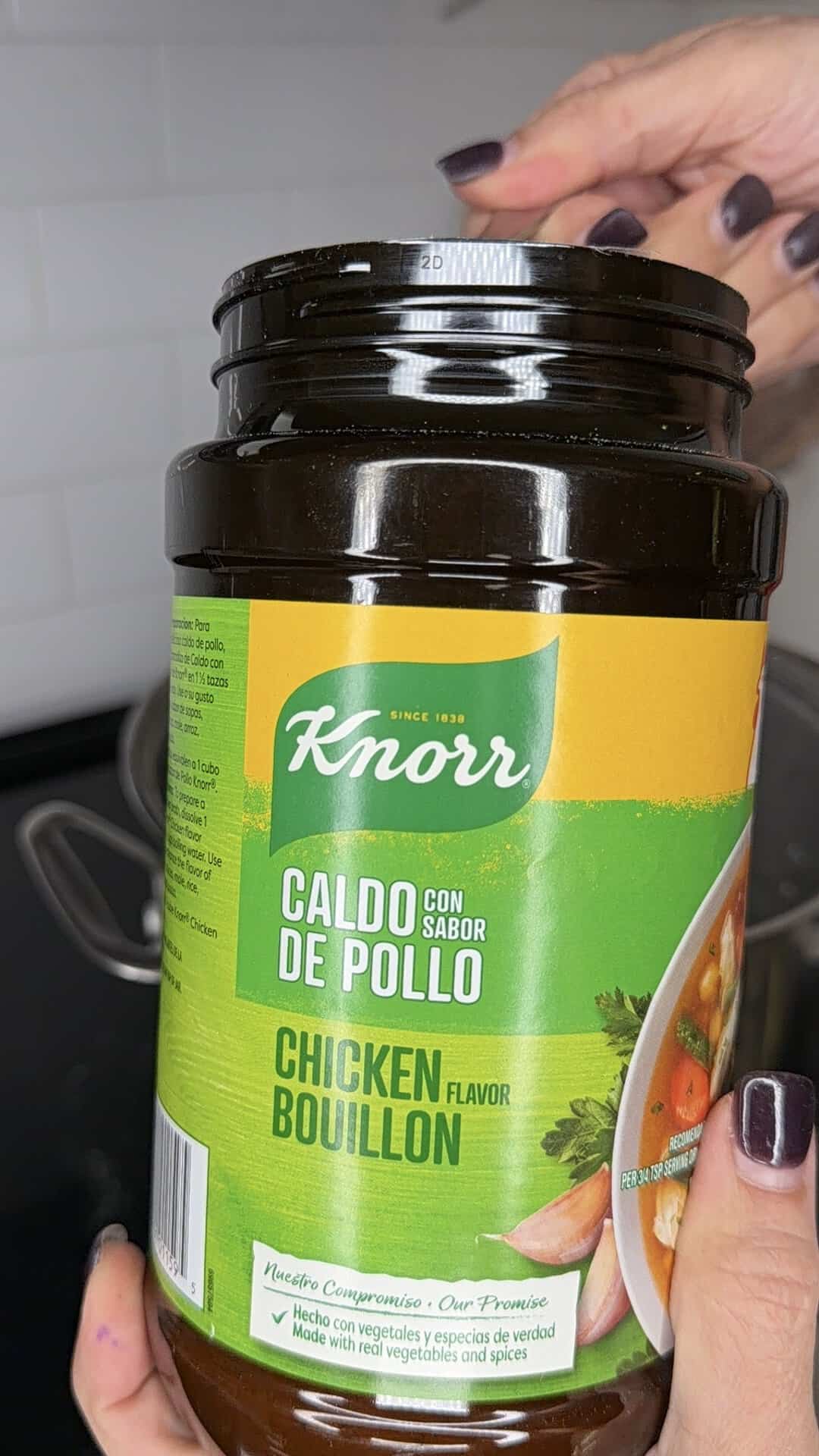 A hand holds an open jar of Knorr Chicken Flavor Bouillon (Caldo con Sabor de Pollo) over a stovetop, with a partial view of a pot and white tiled backsplash in the background.