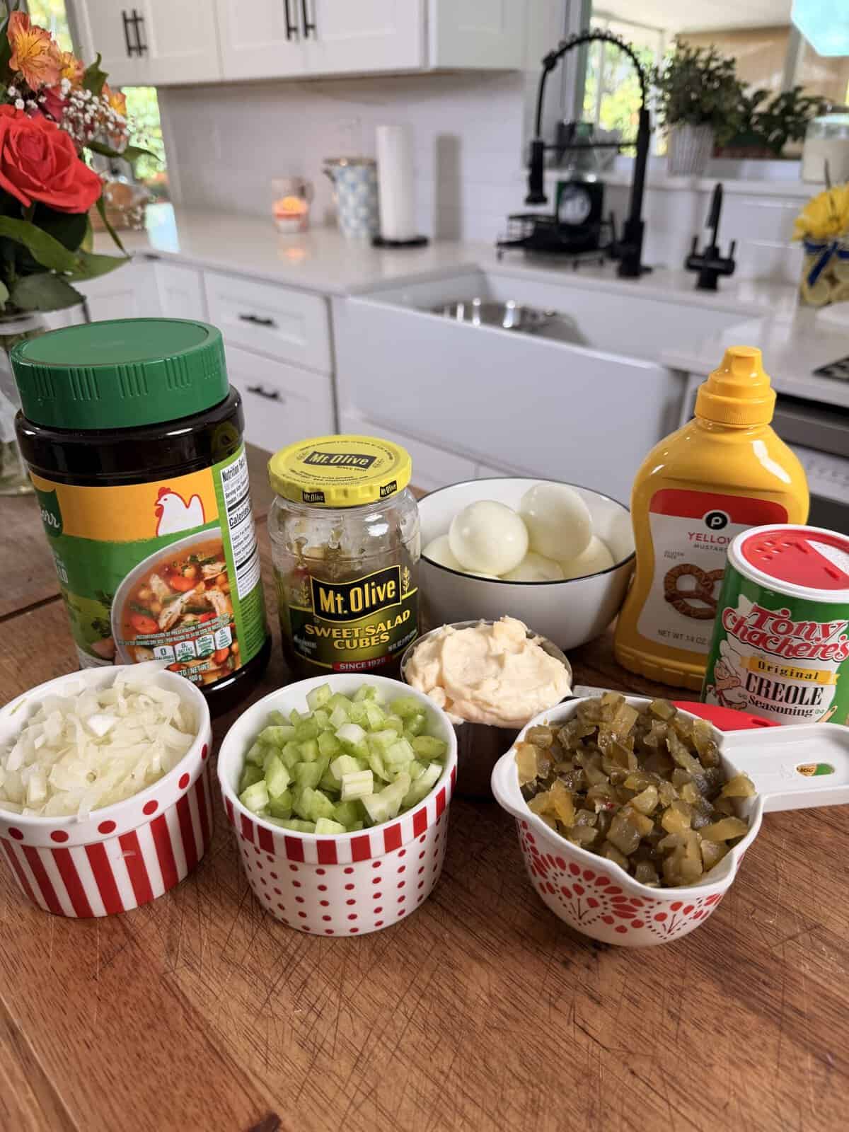 A variety of ingredients for potato salad are arranged on a kitchen counter, including mayonnaise, mustard, boiled eggs, chopped onions, celery, sweet relish, salad cubes, and Creole seasoning.