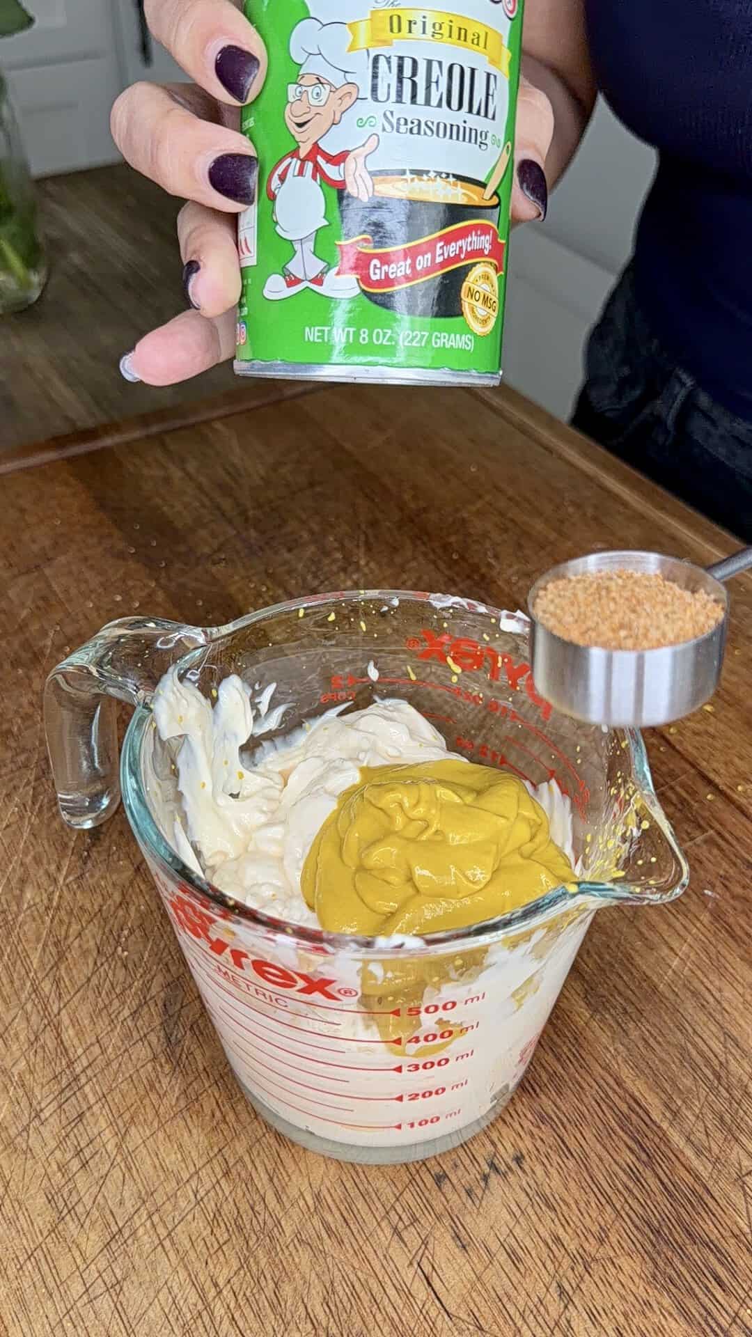 A hand holds a can of Creole seasoning above a measuring cup containing mayonnaise and mustard. A tablespoon of brown seasoning is about to be added to the mixture on a wooden countertop.