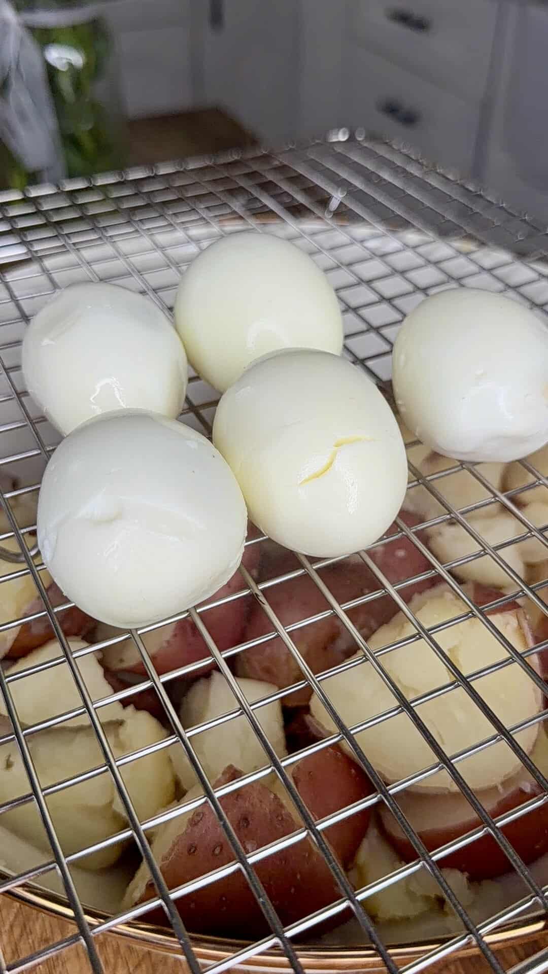 Six peeled hard-boiled eggs rest on a metal cooling rack above quartered boiled red potatoes in a kitchen setting.