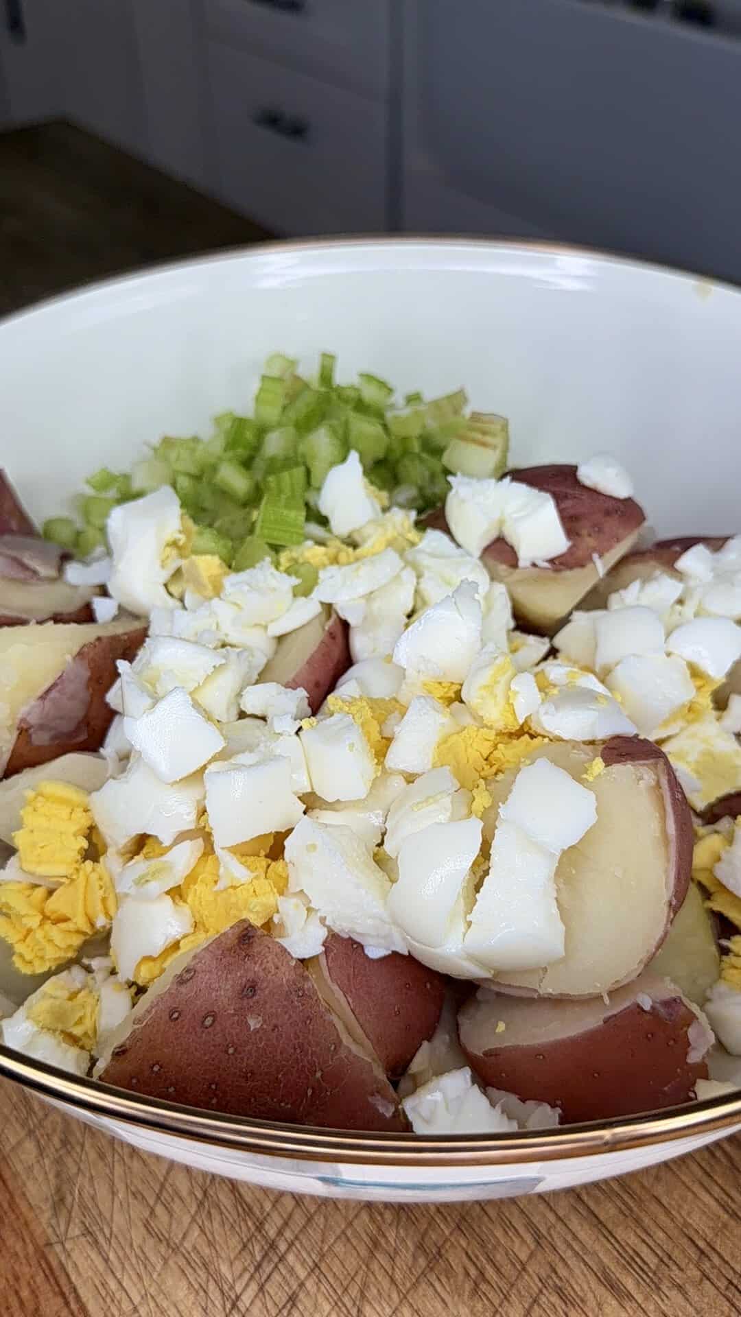 A bowl filled with chopped red potatoes, diced hard-boiled eggs, and sliced celery on a wooden surface, ready to be mixed for potato salad.