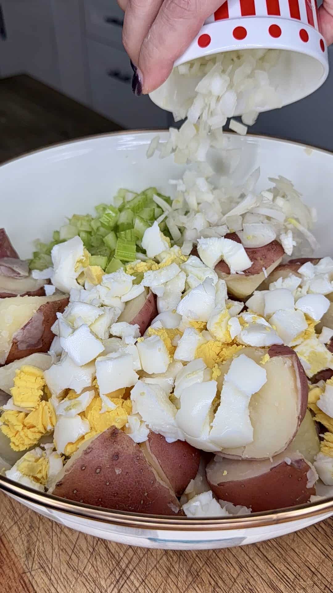 A hand pours chopped onions into a bowl filled with sliced red potatoes, chopped hard-boiled eggs, and celery, preparing ingredients for a potato salad.