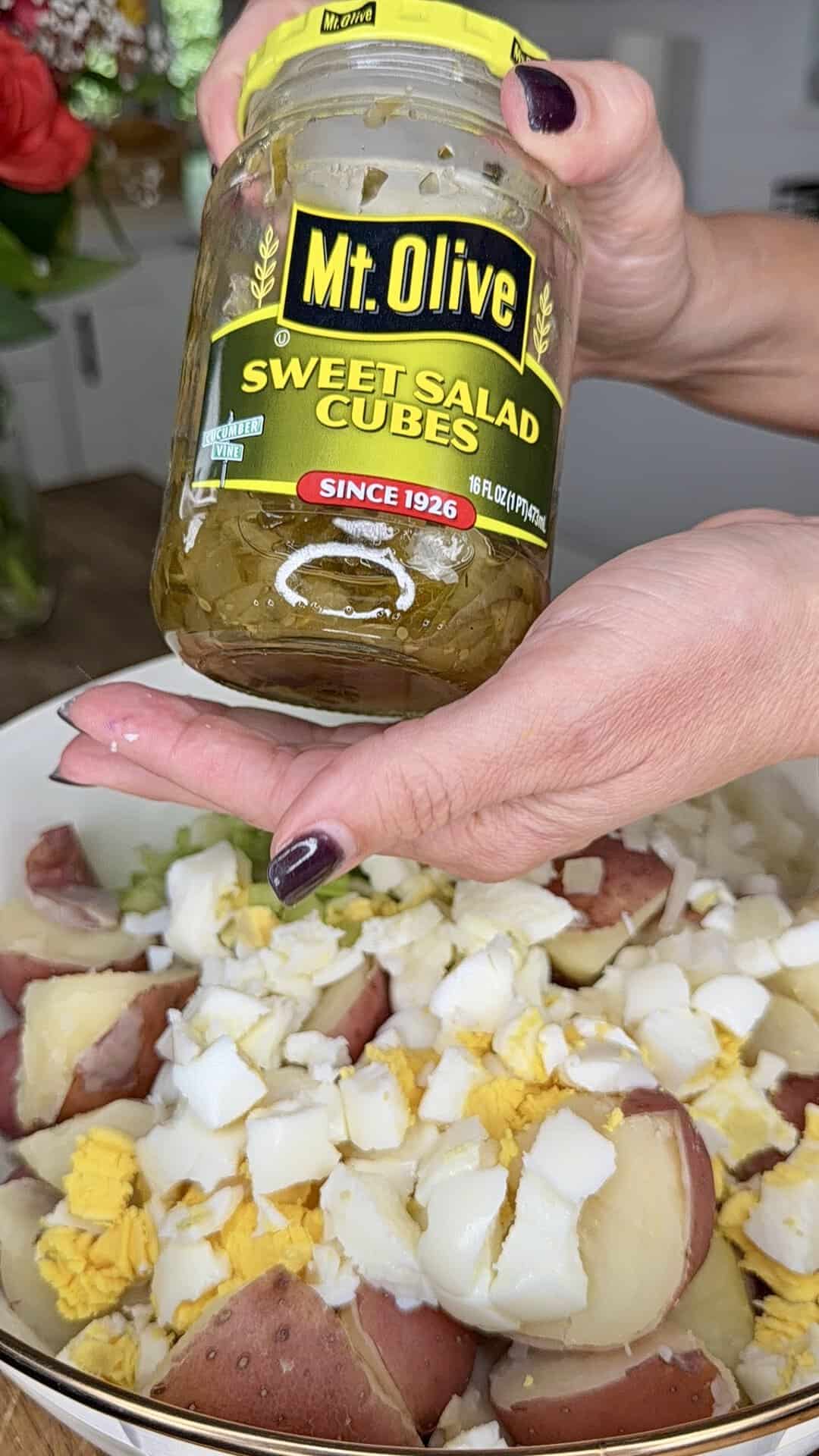 A person holds a jar of Mt. Olive Sweet Salad Cubes above a bowl containing chopped hard-boiled eggs and red potatoes, preparing ingredients for a salad.