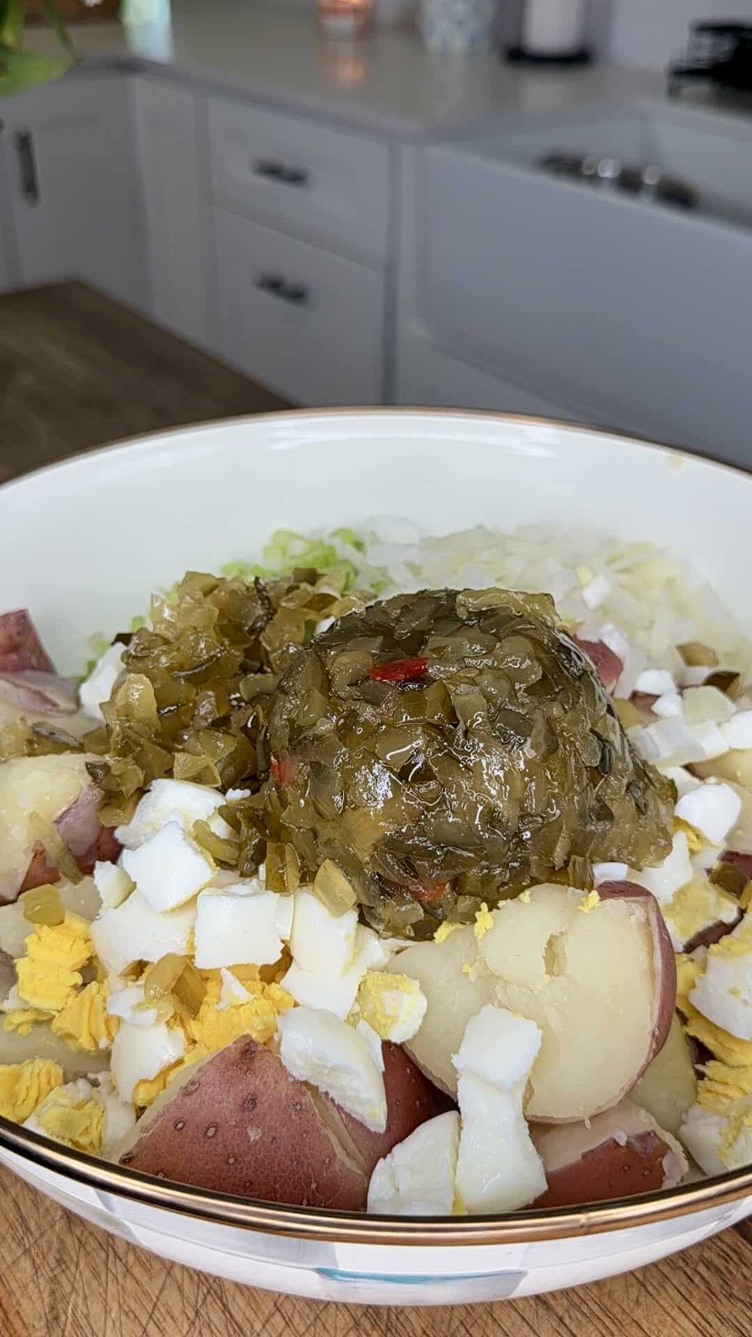 A bowl containing diced hard-boiled eggs, chopped onions, celery, cooked red potatoes, and a large spoonful of relish, sits on a kitchen counter with a sink and cabinets in the background.
