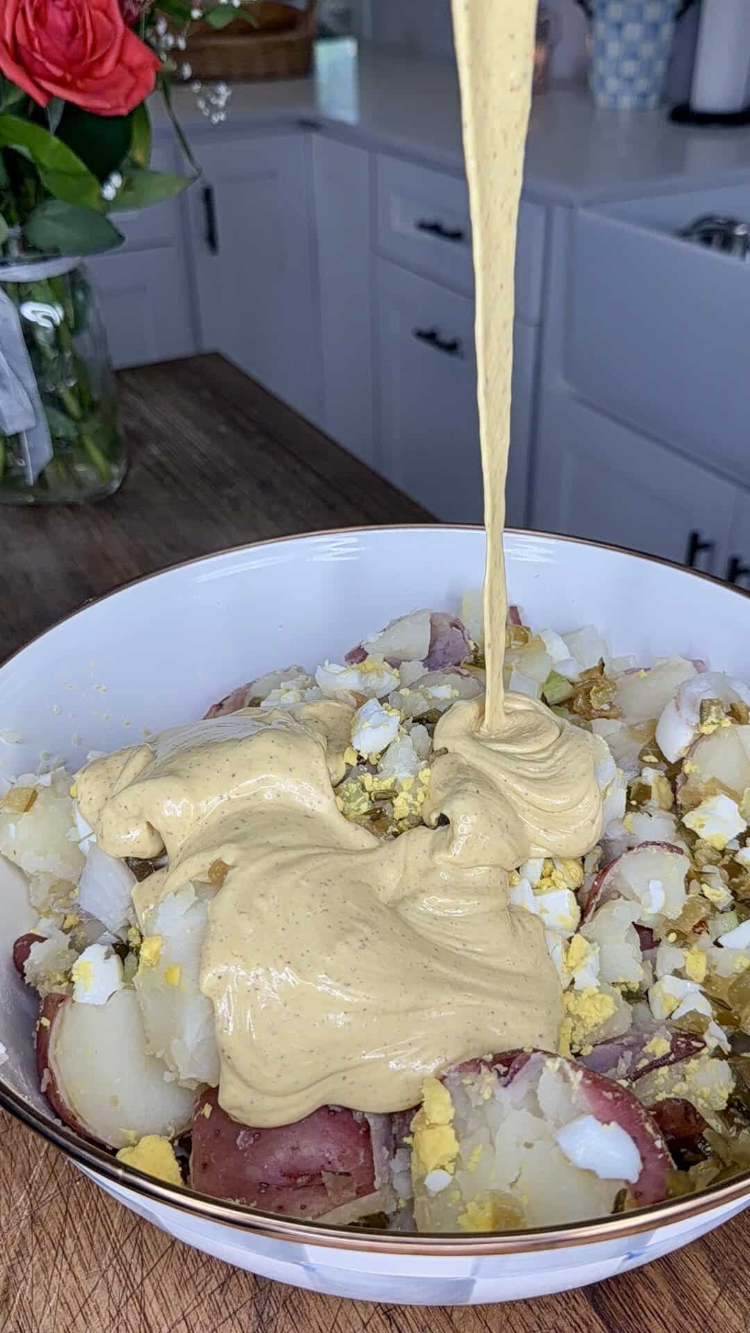 A bowl of chopped boiled potatoes and eggs is being topped with a generous pour of creamy yellow dressing in a kitchen setting. Fresh flowers and white cabinets are visible in the background.