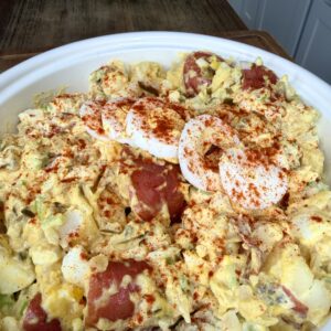 A large bowl of creamy potato salad with chopped red potatoes, sliced hard-boiled eggs, and a sprinkle of paprika, sitting on a kitchen table.