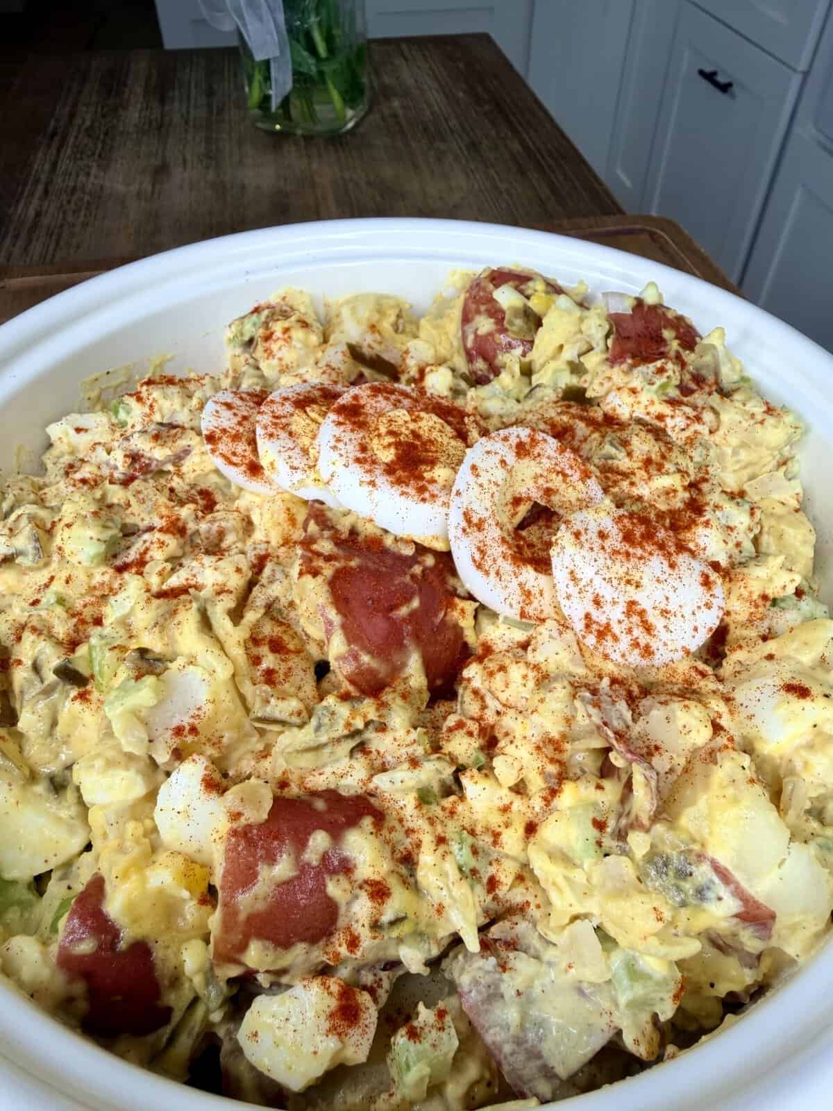 A large bowl of creamy potato salad with chopped red potatoes, sliced hard-boiled eggs, and a sprinkle of paprika, sitting on a kitchen table.