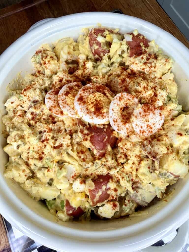 A bowl of potato salad with chopped potatoes, creamy dressing, and sliced hard-boiled eggs on top, sprinkled with paprika for garnish. The bowl is placed on a wooden surface.