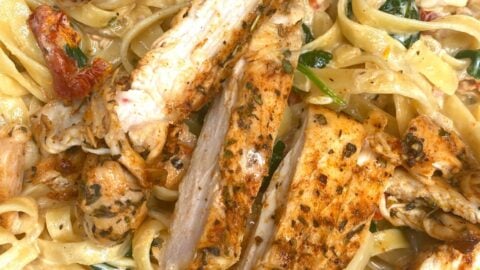 Close-up of creamy pasta with fettuccine noodles, topped with sliced, seasoned grilled chicken, sun-dried tomatoes, and fresh spinach in a rich, creamy sauce.