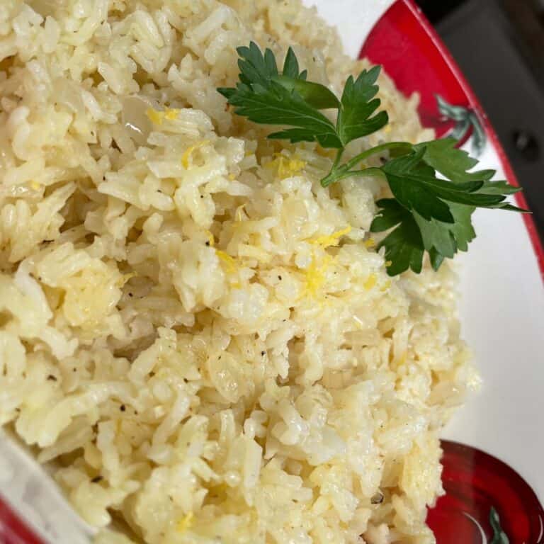 A close-up of a bowl of cooked rice garnished with fresh parsley, served in a red and white dish. The rice appears fluffy and lightly seasoned.