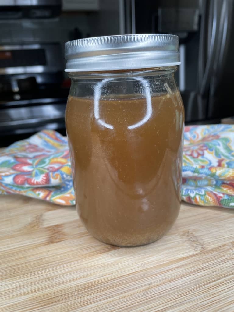 A glass mason jar filled with brown liquid sits on a wooden surface, with a colorful cloth and a kitchen setting in the background.