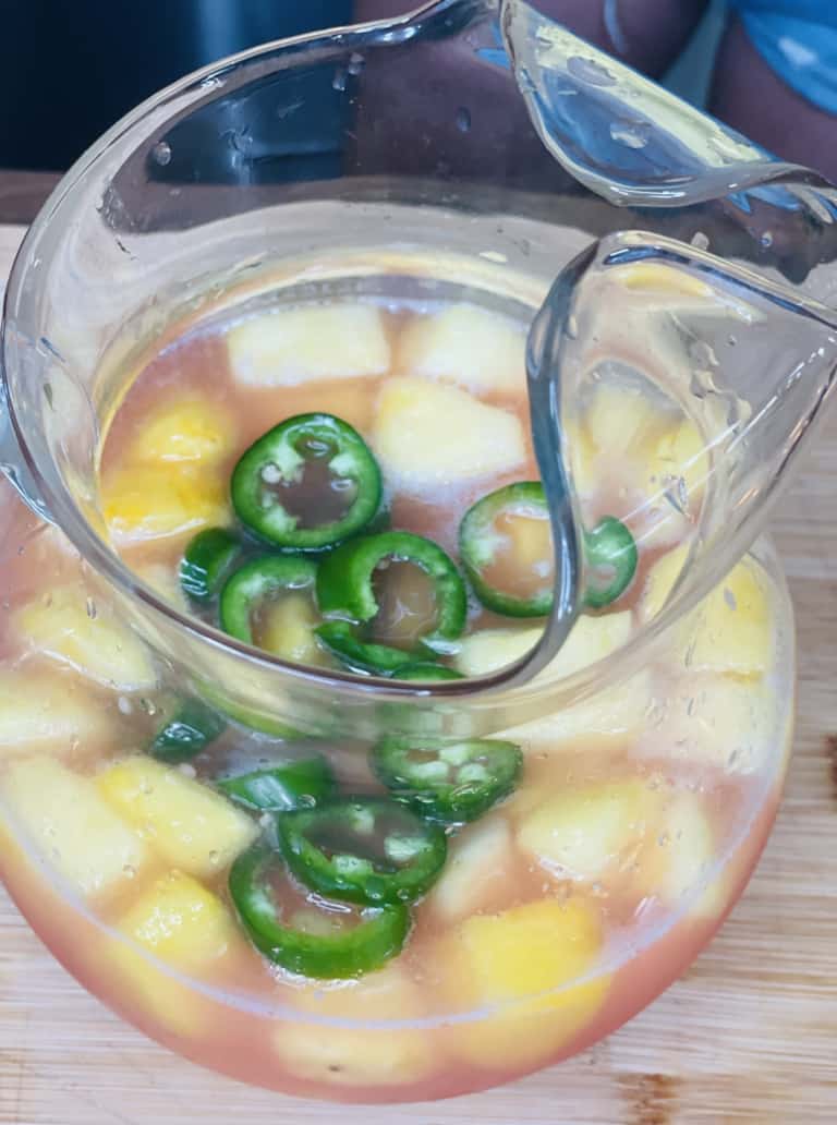A glass pitcher filled with a pinkish drink, ice cubes, pineapple chunks, and sliced jalapeños, sitting on a wooden surface.