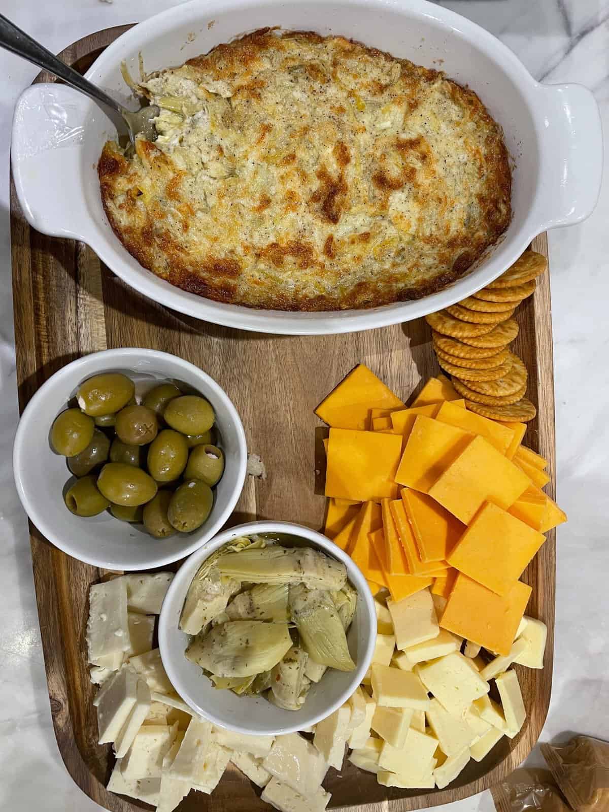 A wooden board with a baked dip in a white dish, a bowl of green olives, a bowl of marinated artichoke hearts, sliced cheddar and white cheese, and round crackers arranged neatly.