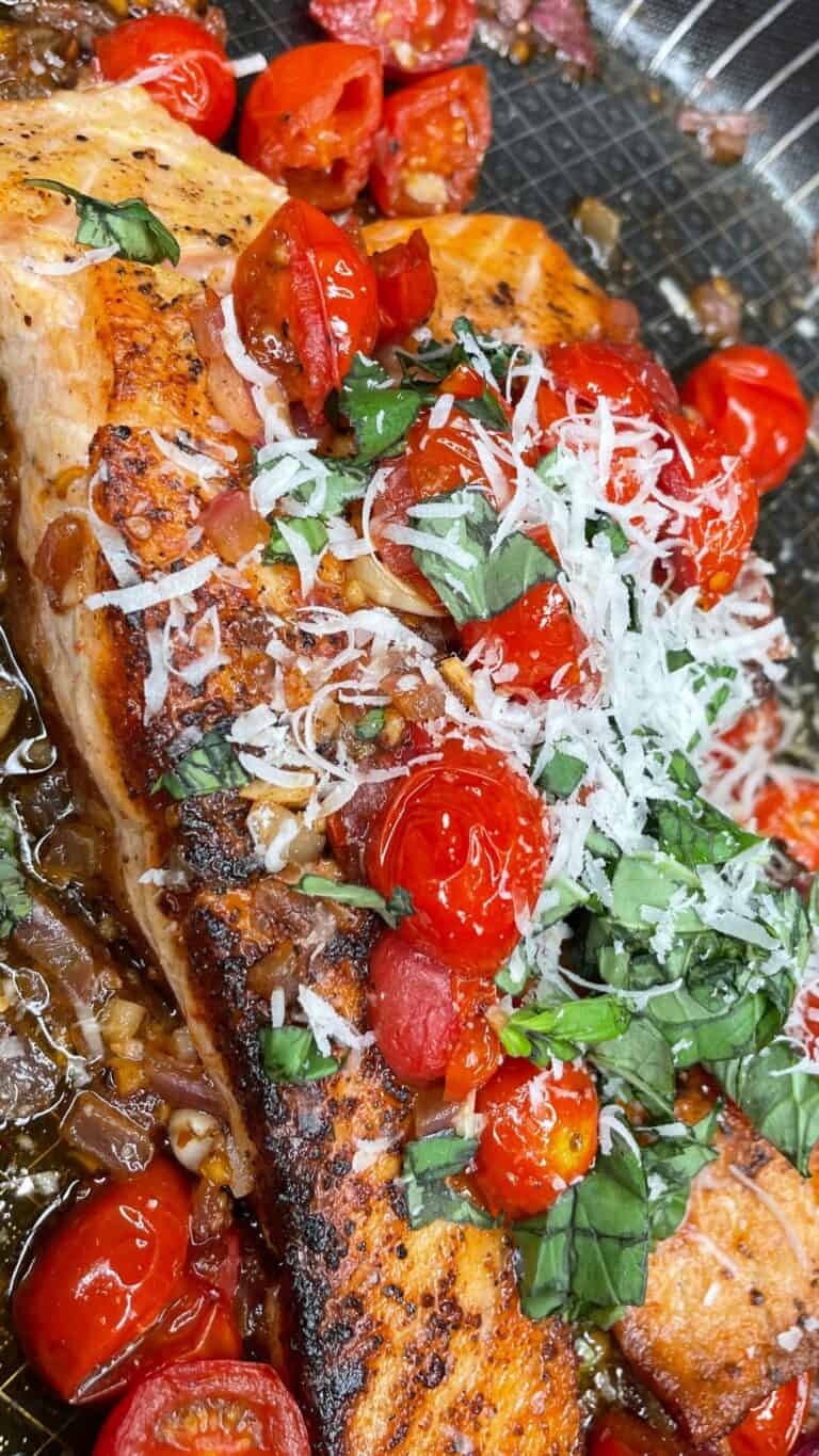 Close-up of a pan-seared salmon fillet topped with sautéed cherry tomatoes, grated cheese, and fresh chopped basil, all in a skillet with visible pan marks and juices.