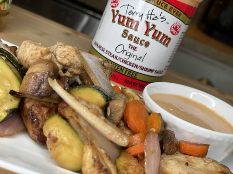 A close-up of grilled chicken and vegetables on a white plate, served with a small bowl of dipping sauce, with a bottle of Terry Hos Yum Yum Sauce in the background.