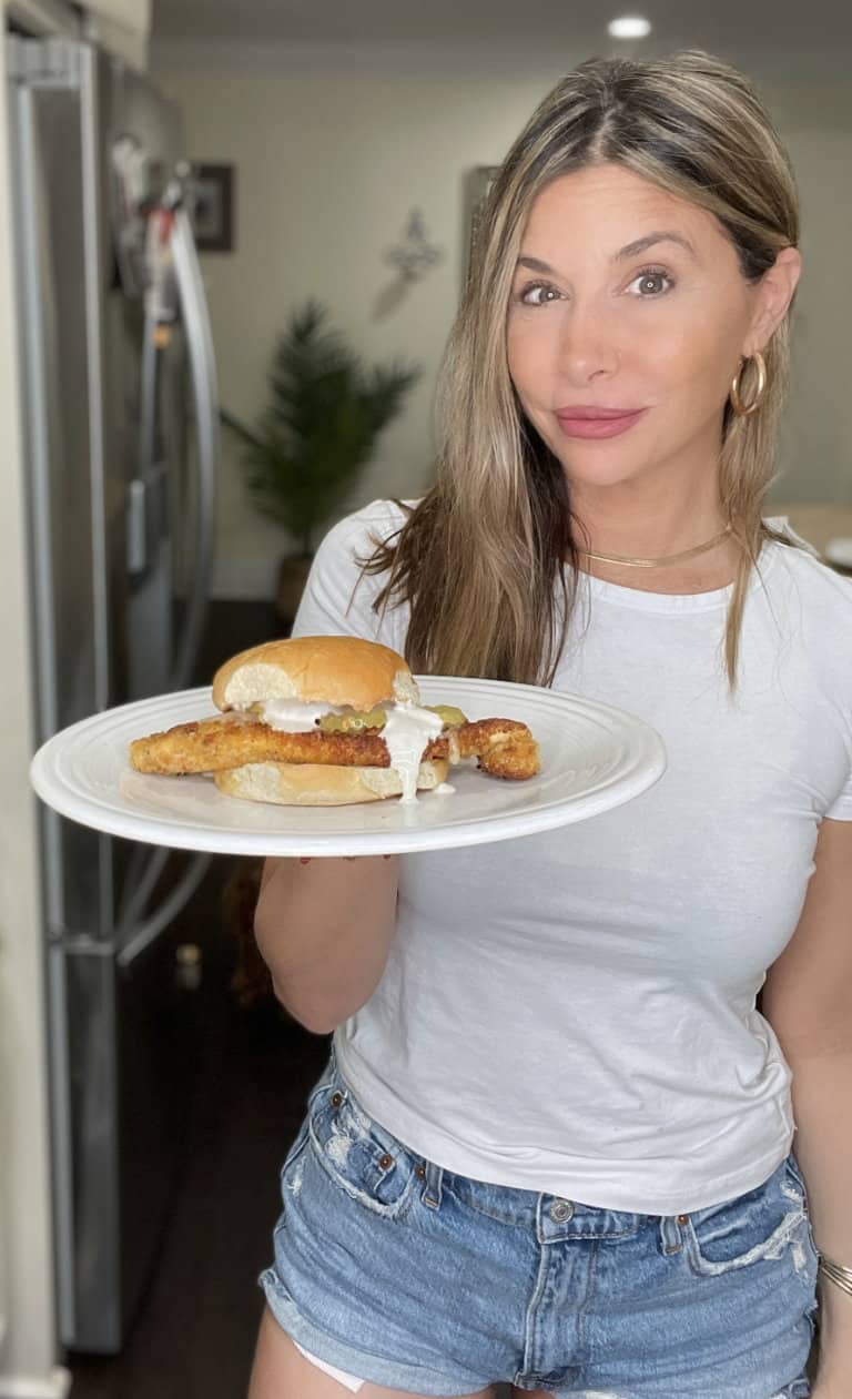 A woman in a white t-shirt and denim shorts holds a plate with a chicken sandwich and white sauce, standing in a kitchen with a refrigerator in the background.