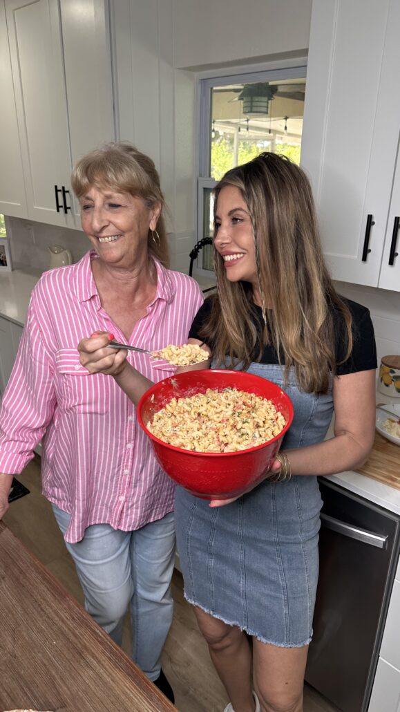 Two women stand in a kitchen smiling. One holds a large red bowl filled with pasta salad and a serving spoon, while the other looks on happily. White cabinets and a window are visible in the background.