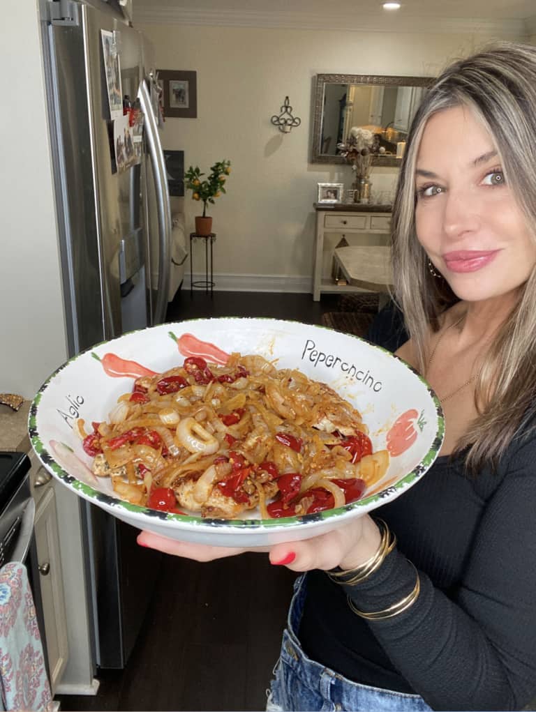 A woman with long brown hair in a black top holds a large bowl of pasta with onions, tomatoes, and peppers in a kitchen, smiling at the camera.