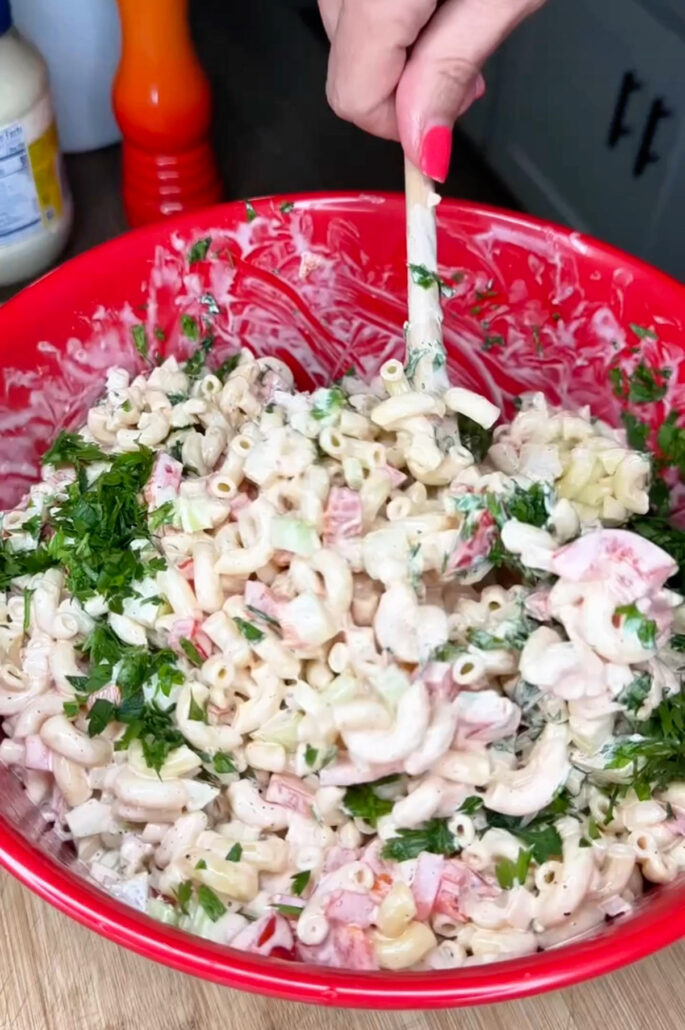 A hand with red nail polish stirs a creamy macaroni salad with parsley, diced tomatoes, and other vegetables in a large red mixing bowl.