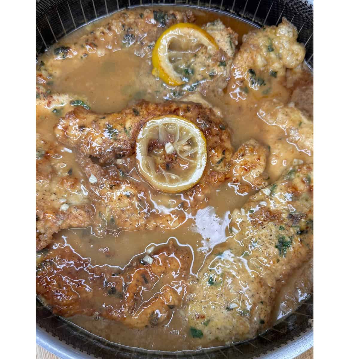 Breaded chicken cutlets in a lemony brown sauce, garnished with slices of lemon and chopped herbs, shown in a pan.