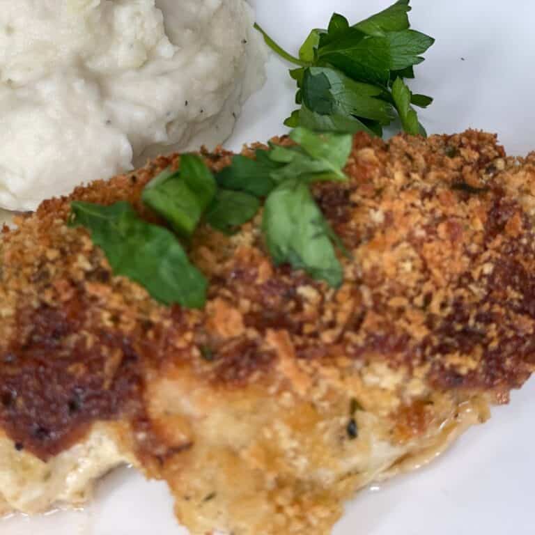 A breaded and baked chicken breast topped with chopped parsley, served with a side of mashed potatoes and a sprig of fresh parsley on a white plate.