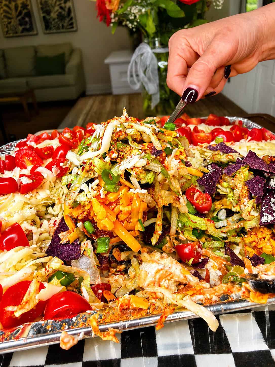 A close-up of a colorful layered salad with shredded lettuce, tomatoes, cheese, crushed chips, and various toppings on a tray. A hand with dark nail polish is serving a portion using a utensil. Flowers and a sofa are in the background.