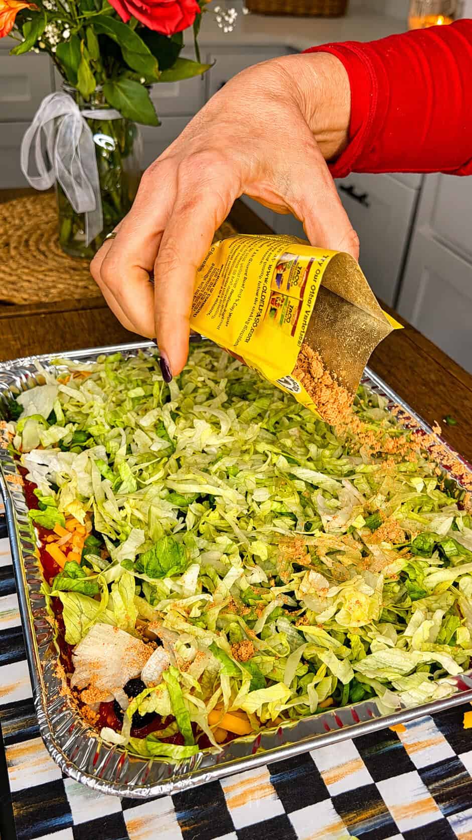 A hand pours taco seasoning from a yellow packet over a tray of shredded lettuce, cheese, and other taco salad ingredients on a kitchen counter.