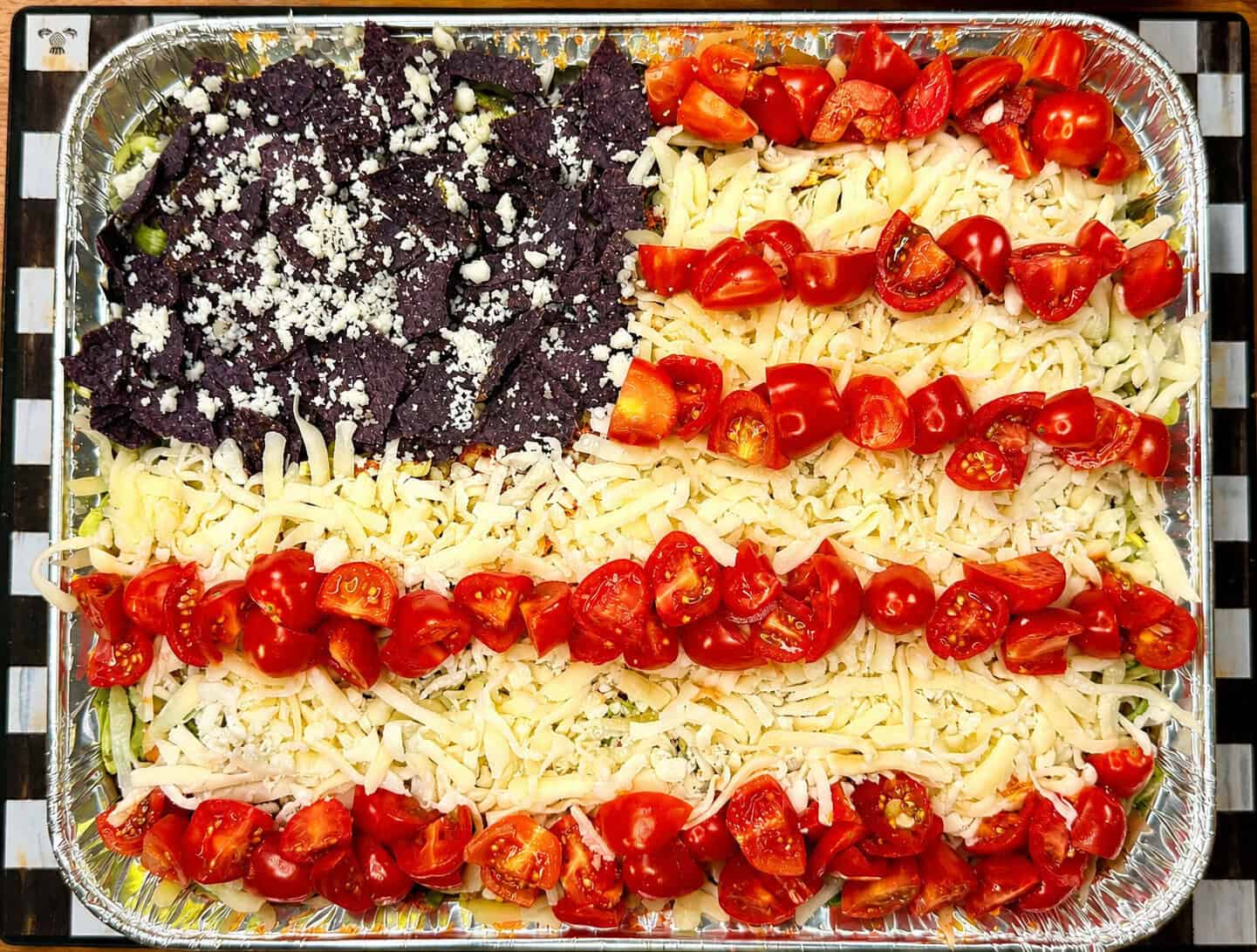 A rectangular tray of dip is decorated to resemble the American flag, with blue corn chips topped with cheese in the top left corner and rows of shredded cheese and chopped tomatoes forming the stripes.